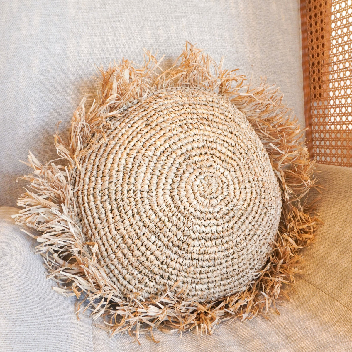 Rundes Soeji Raffia-Dekokissen TARAKAN mit Fransenrand auf hellem Polster, schräg von oben